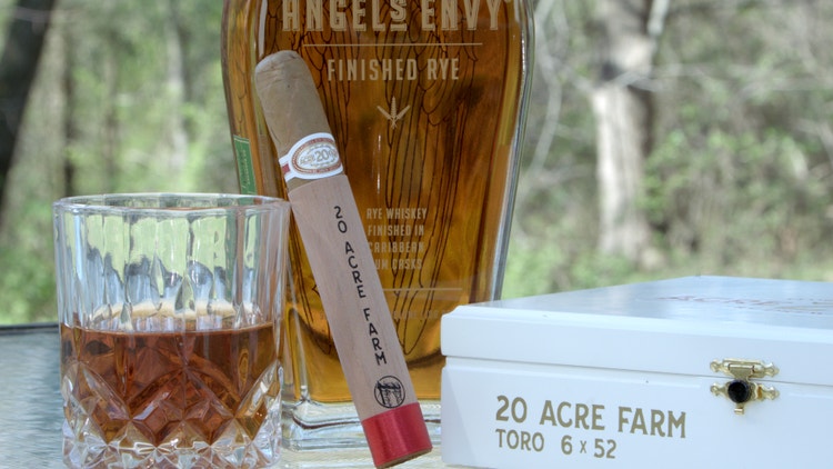 cigar advisor #nowsmoking cigar review 20 acre farm drew estate - cigar leaning against bottle of whiskey glass, bottle of angel's envy finished rye with rocks glass and box in the foreground