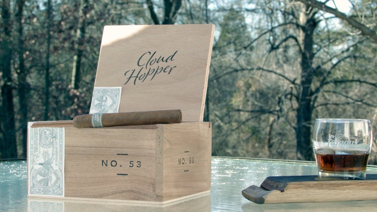 cigar advisor #nowsmoking cigar review video of cloud hopper by warped cigars - setup shot with box and cigar on a table, woodlands in the background, and a whiskey glass