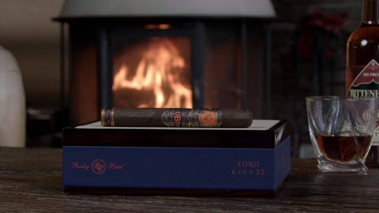 cigar advisor #nowsmoking cigar review rocky patel dbs - setup shot of the cigar on its box with whiskey and a fireplace in the background