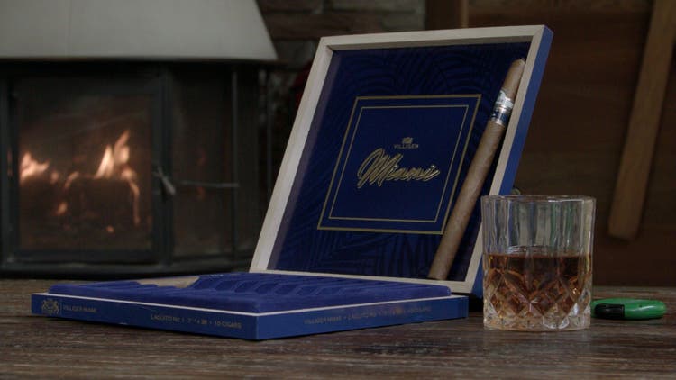 cigar advisor #nowsmoking cigar review villiger miami 2022 - setup shot of cigar resting on its box with a glass of spirits and a fireplace in the background