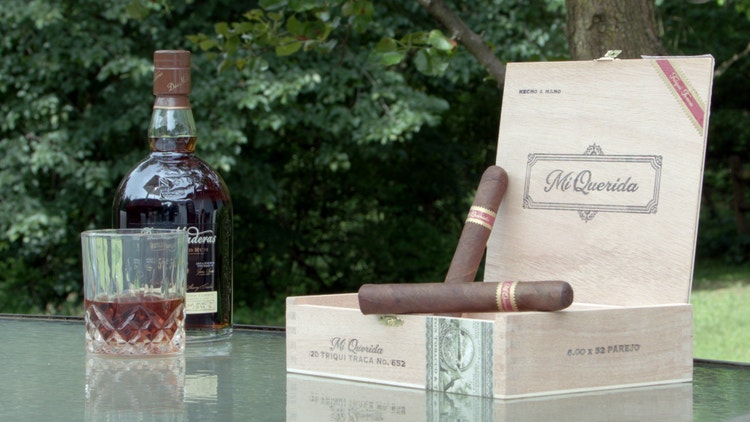 cigar advisor #nowsmoking cigar review dunbarton tobacco & trust mi querida triqui traca - drink pairing with bottle of spirits and glass in the background, cigars and box in the foreground