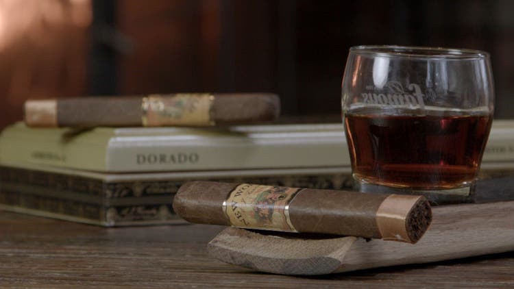 cigar advisor panel review aj fernandez new world dorado - setup shot of cigar box in the background with cigar on a whiskey barrel stave and a glass of bourbon in the foreground